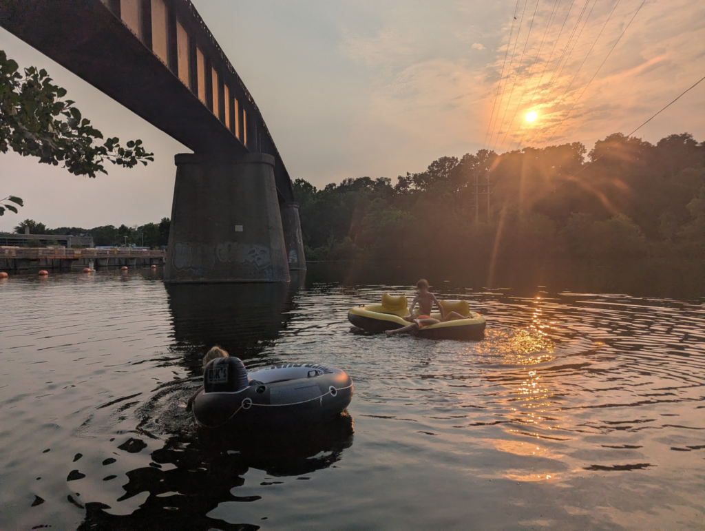 Three kids in two inner tubes, floating on a river near a train bridge at sunset